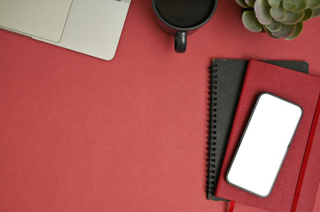Office workspace top view with smartphone white screen mockup, notebook, laptop, coffee cup, potted plant and copy space on red background.の写真素材