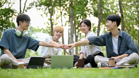 A group of happy, diverse young Asian college students sitting on the grass in the campus park enjoy talking and putting their hands together to cheer each other up. Friendship conceptの写真素材