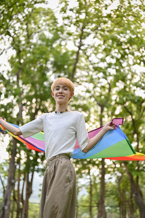 Portrait of an attractive and happy  young Asian gay man in casual clothes with an LGBT rainbow flag standing in the park. Pride parade, LGBTQ+の写真素材