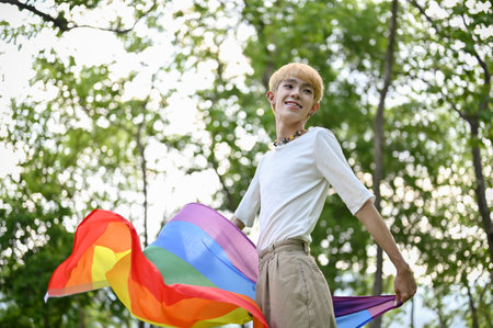 Portrait of an attractive and carefree young Asian gay man with an LGBT rainbow flag standing in the green park. LGBTQ+, Pride monthの写真素材