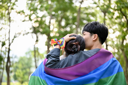 Back view of a happy young Asian man kissing the forehead of his girlfriend and gently hugging her with the LGBT rainbow flag outdoors. Diversity, Couple, Togethernessの写真素材