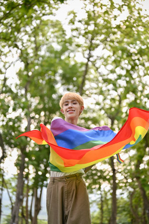 Portrait of an attractive and joyful young Asian gay man with an LGBT rainbow flag standing in the green park. LGBTQ+ conceptの写真素材