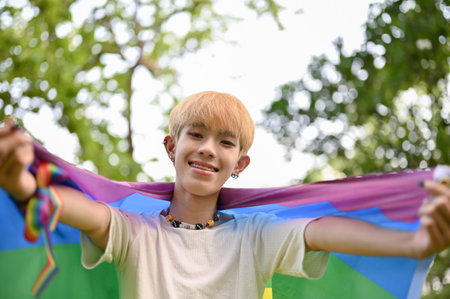 Portrait of an attractive and smiling young Asian gay man with an LGBT rainbow flag standing in the green park. LGBTQ+ conceptの写真素材