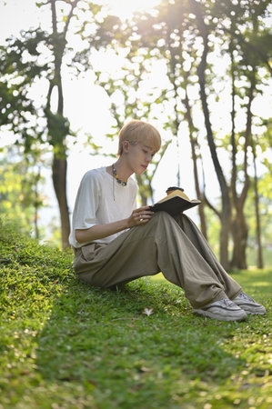 Portrait of a young Asian gay man focuses on reading a book while relaxing in the green park alone on a sunny day. LGBTQ and lifestyle conceptsの写真素材
