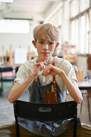 A portrait of an attractive and confident young Asian queer or gay man artist in an apron sitting on a chair in his creative studio. LGBTQ+ and creative work conceptの写真素材