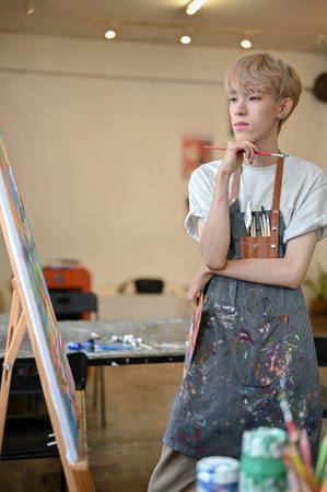 Portrait of a professional and talented young Asian queer artist in an apron looking at his picture on a canvas easel, focuses on painting in his studio.の写真素材