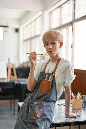 Portrait of a creative and confident young Asian gay artist in a dirty apron looking at the camera while standing in his creative studio. LGBTQ+ and creative job conceptsの写真素材