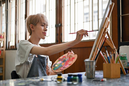 A creative and talented young Asian gay man or queer art student is painting watercolors on a small canvas at his desk in the studio. LGBTQ+ and creative job conceptsの写真素材