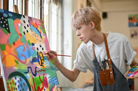 A professional and talented young Asian queer artist in an apron focuses on painting his picture on a canvas easel in his studio.の写真素材