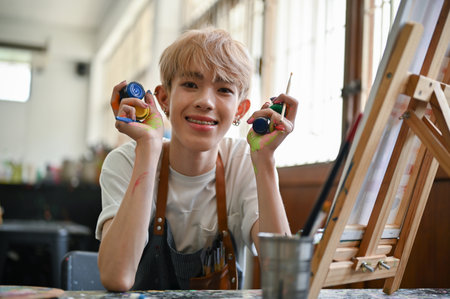 A happy and talented young Asian gay artist or painter sits at his workspace with a canvas on a table and painting equipment in his hands. LGBTQ+ and hobby conceptsの写真素材