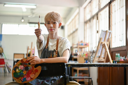 A professional and talented young Asian gay man or queer artist sits on a chair in his creative studio with a paintbrush and art palette in his hands.の写真素材