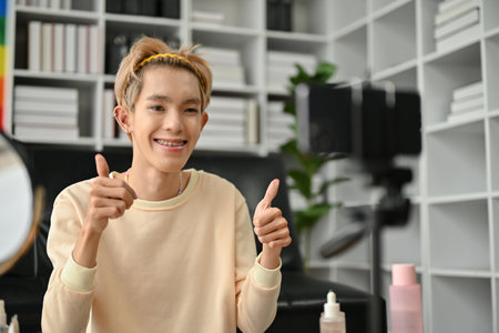 A happy and smiling young Asian gay man blogger is showing his thumbs up to the camera and recording his makeup tutorial video in his room. LGBTQ+ conceptの写真素材