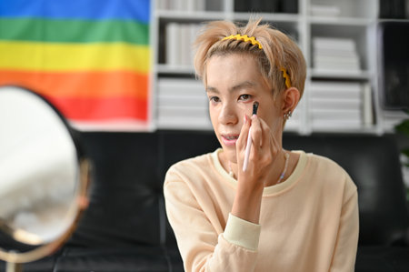 A young Asian queer man looking in the mirror and applying foundation make-up at home. LGBTQ+ lifestyle conceptの写真素材