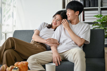 A cute and lovely young Asian couple is hugging while enjoying watching TV on a couch in the cozy living room together. love and relationship conceptsの写真素材