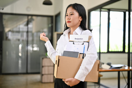 Confident young Asian female office worker feels happy to quit her job, holding a cardboard box with her resignation letter.の写真素材