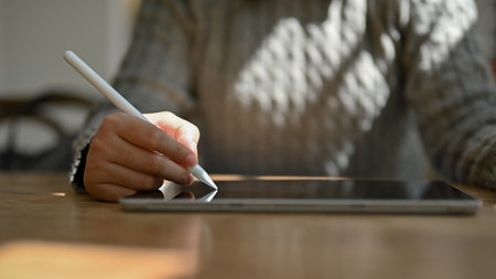 Close-up image of a female in a cozy grey sweater using a stylus pen to draw or write on her digital tablet while sitting in a cozy coffee shop.の写真素材