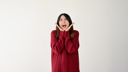 Pretty and excited young Asian woman in a cozy red sweater is looking up, hands on her chin, and saying wow. isolated on white backgroundの写真素材