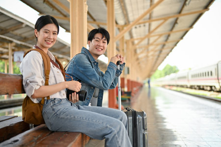 A happy and lovely young Asian couple with their suitcase is traveling together, waiting for their train on the platform at the railway station.の写真素材