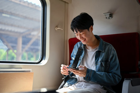 A happy and handsome young Asian male traveler backpacker using his smartphone while sitting in his seat on a train. Technology and transportation conceptsの写真素材