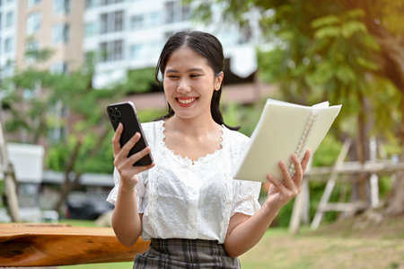 A beautiful and happy young Asian female is holding a book and looking down at her smartphone screen...