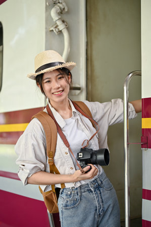 A beautiful and cheerful young Asian female tourist with a straw hat, camera, and backpack is on a train ladder, arriving at her destination station. Solo traveling conceptの写真素材