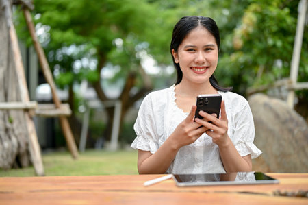A smiling and beautiful Asian woman is looking at the camera and holding her smartphone while sitting at a table outdoors.の写真素材