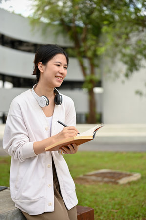 A portrait of a beautiful and happy young Asian female college student in a comfy cardigan sits at a table in the campus park, writes her diary, or makes notes of her creative ideas.の写真素材