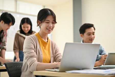 Close-up image of a beautiful and smart young Asian female worker in a startup team is working on her tasks on hera laptop while her colleagues are in the room.の写真素材
