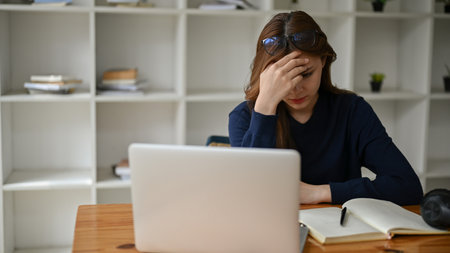 A stressed and thoughtful young Asian businesswoman sits at her desk in the office, trying to figure...