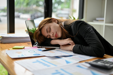A tired and sleepy young Asian businesswoman or female office worker falls asleep at her office desk with a bunch of reports on the table. overworked, exhausted, lazy, long day at workの写真素材