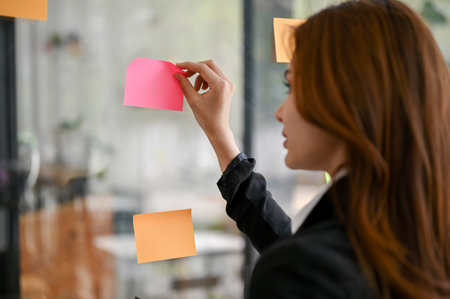 Close-up image of a beautiful young Asian female marketing assistant looking at the sticky note on a glass wall. ideas, notes, memo boardの写真素材