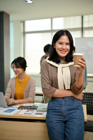 A portrait of an attractive young Asian female graphic designer or website developer stands in the office with a coffee cup in her hand while her coworkers are working in the room.の写真素材