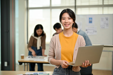 A beautiful young Asian female graphic designer or website developer stands in the co-working office with a laptop in her hand while her coworkers are working together in the room.の写真素材