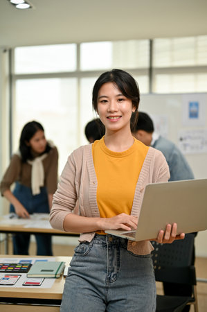 A portrait of a beautiful and charming young Asian female graphic designer or website developer stands in the co-working office with a laptop in her hand.の写真素材