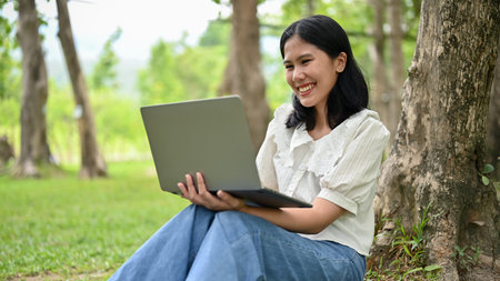 A happy young Asian female in casual clothes is watching some online content on her laptop while...