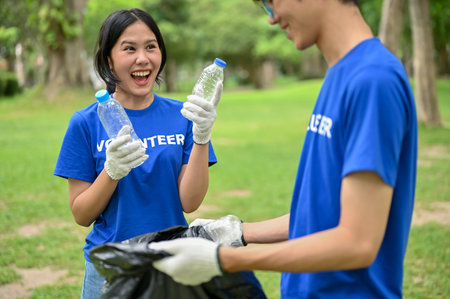 A joyful and happy young Asian female volunteer collecting trash and plastic bottles into a garbage...