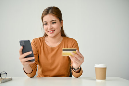 An attractive Asian woman sits at a table and connected her credit card to a shopping app through her smartphone, enjoys shopping online at home.の写真素材