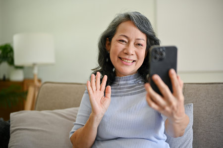 An attractive and happy mature middle-aged Asian woman is on a video call with her grandchild while...