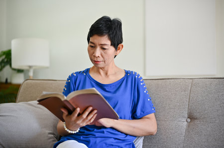 A focused mature middle-aged Asian woman is reading a book on a sofa in her living room. retirement lifestyleの写真素材