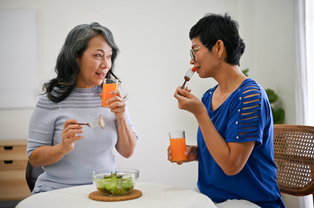 Two lovely and happy retired Asian female close friends are enjoying their healthy brunch and chatting in the dining room, spending time on the weekend together.の写真素材