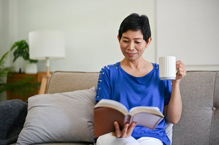 A relaxed retired Asian woman in comfy clothes enjoys her morning coffee and reads a book on a sofa...
