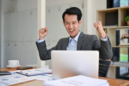 A cheerful millennial Asian businessman in a formal business suit is raising his hands, looking at his laptop screen, and celebrating his success in the office. project approved, getting job promotionの写真素材