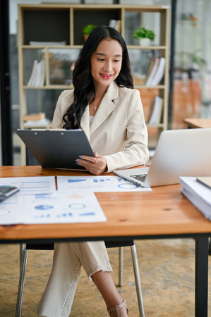 A portrait of a beautiful millennial Asian businesswoman or female executive manager in a trendy suit focuses on business reports and working on her tasks on her laptop at her desk in the office.の写真素材