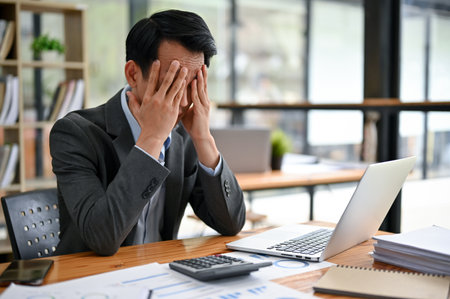 A stressed and frustrated millennial Asian businessman is covering his face, pensively making a plan to solve the problems, upset with his project failure, sitting at his desk in the office.の写真素材