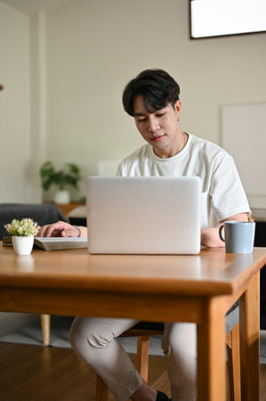 A portrait of a handsome young Asian man using his laptop at a table in a minimalist living room.の写真素材