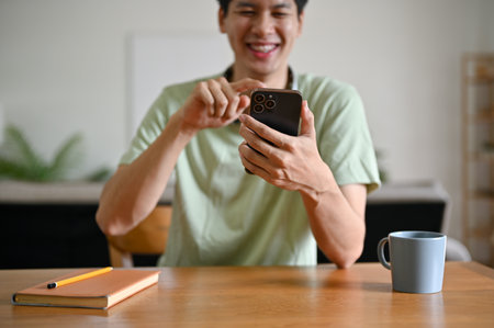 A cheerful young Asian man is using his smartphone, scrolling on social media, chatting with his friends, or enjoying online shopping on the app while sitting at a table in the living room.の写真素材