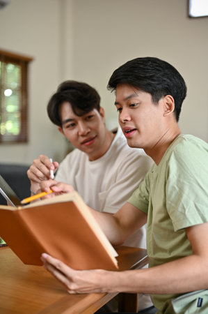 A smart young Asian male college student is tutoring and explaining something in a book to his friend while studying at home together.の写真素材