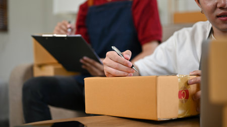 A handsome young Asian gay man writing a customer's address on a cardboard box and preparing...