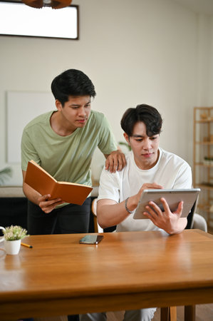 Two focused young Asian male college students do their homework at home together, searching for...