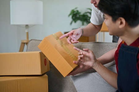 A happy young Asian male online seller checking the customer's address on a cardboard box and preparing shipping products with his boyfriend at home. LGBTQ+, small business, e-commerceの写真素材